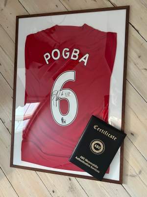 Paul Pogba signerad matchtröja från säsongen 16/17. Paul Pogba gjorde totalt 157 tävlingsmatcher för Manchester United mellan åren 2011-2012 samt 2016-2022. På andra bilden visas ett äkthetsbevis för att aurografen är skriven av Pogba och vilket datum.