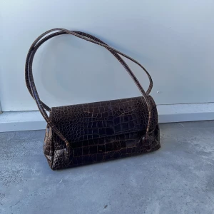 VINTAGE VÄSKA - Brun vintageväska med krokodilmönster. Tre innerfickor, en med dragkedja. Nyckelring i väskan. Även en ficka med dragkedja på utsidan (baksidan). Superrymlig med långa axelband. Inga skavanker eller dylikt.   Pris: 100 kr, kan fraktas för 69 kr (spårbart)