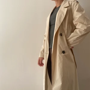 Beige trench coat - Lång trench coat med blårutig bakdel. Jag är 175 lång och den går ca 1 dm under knäna på mig. Köptes i barcelona så minns inte vad butiken hette. I skick som ny!