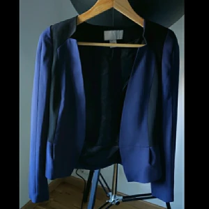 Kavaj med volang - Unik marinblå kavaj med svarta partier och volang i ryggänden från HM, strl 36. Fin till fest och jobb. Använd 1 gång. Skriv i chatt vid intresse. Möts upp i Hägersten. 