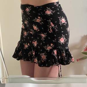 Svart kjol med rosa blommor. Dragkedja på sidan av höften med spänne. Snörena på kjolen kan man justera själv och välja hur hög slits man vill ha.