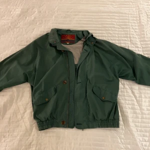 Grön jacka - Grön jacka i ungefär samma material som en vindjacka. Strl M från Redstone. Knappar, dragkedja och framfickor finns. Använd 1 gång.