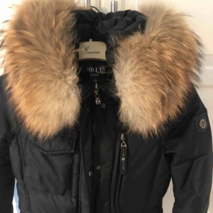  - Figursydd Hollies jacka med avtagbar päls. Jackan är svart med brun päls och fickor framtill. Plagget är använt ungefär tio gånger tidigare men är så gott som nytt. Plagget köptes in för 3000kr
