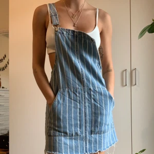 Randig hängselklänning - Blå hängselklänning i jeans med vita tunna ränder. Storlek S. Fickor fram. Bra skick. 