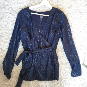  - Säljer en mörkblå blus med glittriga "prickar" på, omlott med band runt midjan. Den är endast provad och är därmed i nyskick (prislapp sitter kvar).