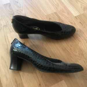 Scholl finskor glänsande svarta - Svarta finskor klackskor i glänsande svart ormskinn (imitation). Står stl 6 1/2 och är då stl 40. Bra kvalitet från Scholl, välbehållna. Gulddetalj bakpå. 
