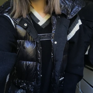 Svart lång väst med huva - Superfin lång väst i svart glansigt material, har bara använt den några enstaka gånger och den värmer helt perfekt på hösten och våren😍