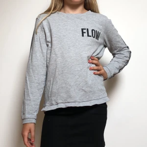 Grå tröja med svarta sträck och tryck - Ljus grå långärmad tröja med svarta sträck på armarna. Sedan står det ”Flow” på framsidan och på baksidan finns ett ”v” format hål.