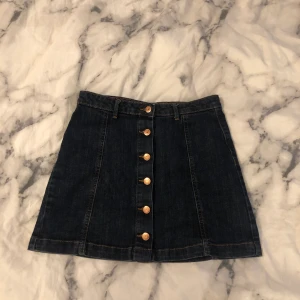 Jeanskjol med knappar strl. 38 - En jeanskjol med knappar från ginatricot i en mörkblå tvätt med kopparfärgade knappar. Lite liten i midjan och runt rumpan för mig (strl M-L och 168 cm). Jätteskönt material och riktigt bra passform! Kan mötas i Stockholm, annars frakt 40 kr :)