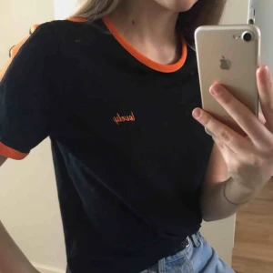  - Svart t-shirt från NA-KD med oranga detaljer. Står lovely på framsidan och har oranga linjer på axlarna (frakt tillkommer). 