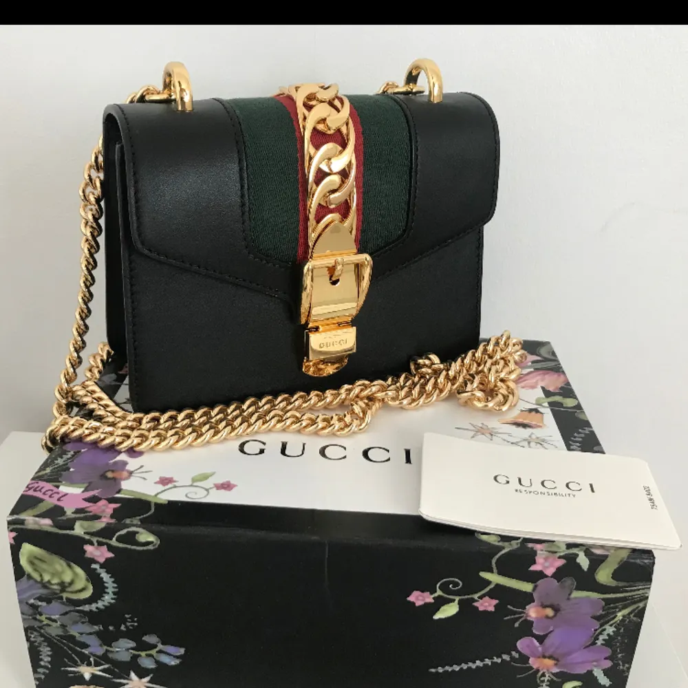 Gucci Sylvie Mini läderväska i svart med guldkedja och en avtagbar web rosett i blått och rött. Tjock guldkedja med spänne som öppnar väskan. Använd fåtal gånger/som ny (säljes pga för lite använd) Orginalpris: €1690 . Accessoarer.
