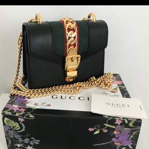 Gucci Sylvie Mini läderväska i svart med guldkedja och en avtagbar web rosett i blått och rött. Tjock guldkedja med spänne som öppnar väskan. Använd fåtal gånger/som ny (säljes pga för lite använd) Orginalpris: €1690 