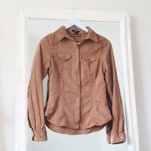 Brun skjorta mocka imitation 36 - Färg: brun.  Strl: 36.   Snygg skjort-blus från H&M, i mockaimitation. Använd vid ett par tillfällen, men i fint begagnat skick. Figursydd och detaljer av fickor framtill. Sluts igen med knappar framtill. Fraktkostnad tillkommer, betalning via swish