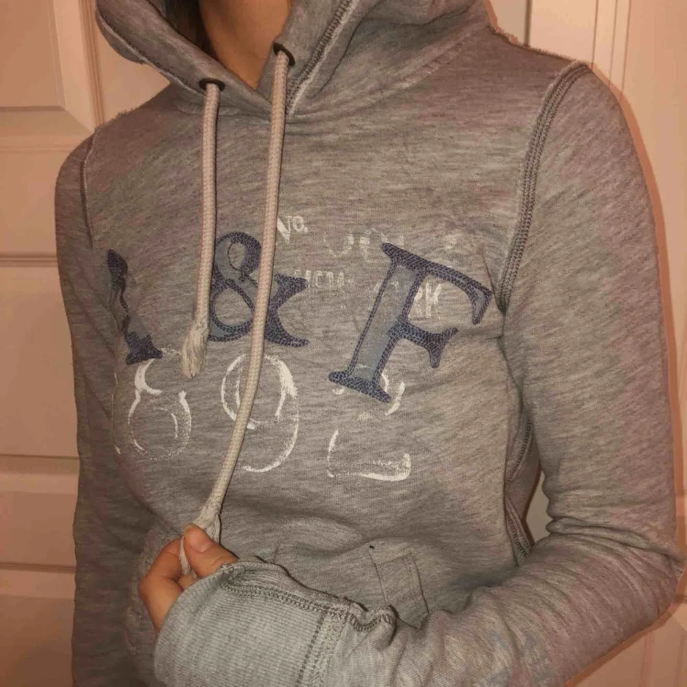En tight A&F hoodie i bra skick. Mellangrå färg, blå text på bröstet och på armen. . Hoodies.