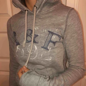  - En tight A&F hoodie i bra skick. Mellangrå färg, blå text på bröstet och på armen. 