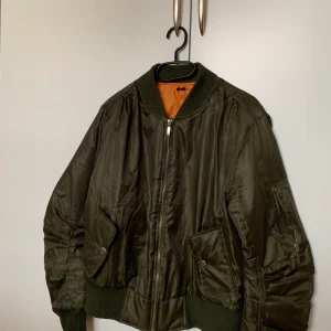 Bomberjacka Grön - Grön bomberjacka strl S, inköpt något år sedan. Märke: Ingen aning. Bra skick, inga slitdetaljer. Orange, kilt-mönstrad insida. Fickor fram.