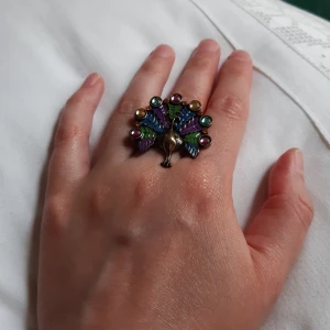 Ring påfågel - Ring som är ställbar till önskad storlek, bronsaktig metall med målade fjädrar och färgade strasstenar. Ca 3,5 cm tvärs över. Pris: 10 kr plus frakt, eller 10 smycken för 50 kr plus frakt.