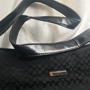 Svart Handväska - Snygg svart handväska köpt second hand. Lite slitage på bandet, annars fint skick. Fack med dragkedja finns på baksidan och även innuti. 30cm bred, 18cm hög. Frakt kostar 44kr