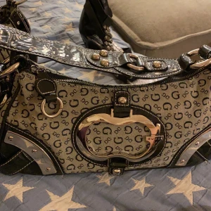 Guess handväska köpt i Guess affär i USA - Har sänkt den från 850kr ordinarie pris ca 500 usd!  Väskan ser svart ut och har detaljer & axelband i lack mörk butelj grön som ser svart ut. Nitar och andra detaljer  i Chrome. 