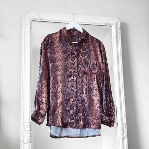 Snake print blus Gina 36 - Färg: brun mönstrad. Strl: 36.  Meterial: 100% polyester. Fin blus från Gina Tricot. Knäpps med knappar. Detalj av en ficka på ena bröstet, samt krage. Skönt mjukt och glansigt material som är skrynkelfritt och har en fin lyster. Längre baktill. Passat både fest och vardag! Använd vid två tillfällen, så gott som ny!!