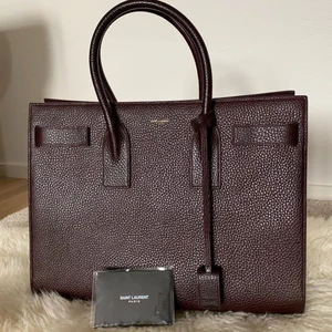 Saint Laurent väska - Selger min burgundy Calfskin-leather large Sac de Jour Tote Bag. Denne vesken har jeg kjøpt fra net-a-porter i 2015. Har brukt den en gang, så man kan si at den er som ny. Fargen passer til alt, og blir fort utsolgt. Den har tre seksjonen en med gull glidelås , en lomme på innsiden, og to nøkler til låsen. H:28cm L:36cm B:14cm Vesken er litt for stor for min livsstil, men den passer veldig bra til jobb med mindre aktivitet og studier pga den har plass til alt og mye mer:)  Nypris. 24.500kr