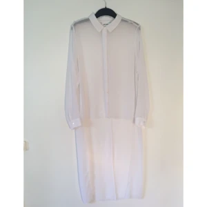  - Vit genomskinlig skjorta, kort fram och lång bak. Genomskinlig plast högst upp på ryggen. Lite trådig i kanterna längst ner, beror på att det inte är några sömmar i kanterna