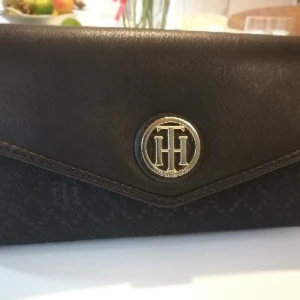  - Tommy Hilfiger plånbok. Väldigt sällsynt i Sverige. Köpt i USA.  Ny, oanvänd med etikett på. Köpt i USA. Färg : svart med detaljer i guldfärg samt gjord i tyg.  Gratis leverans i söderort.  Mått: Höjd : 10 cm Längd/Bredd : 20 cm  Produktinformation: Dragkedja i full längd Fack för kreditkort Myntficka med dragkedja Dragspelsfack
