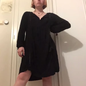 En svart tunikaklänning - En svart tunikaklänning med en brodering med hål i sig. Snören från kragen. Gjord som en L men passar flera storlekar.