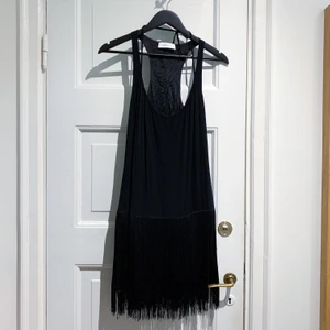 Klänning med fransar - Selected Femme, stl 34. Svart klänning med fransar i Charleston / Great Gatsby stil. Använd en gång. Dragkedja i sidan. Hel och ren. Snören vid ringningen kan justera ringningen. Fransar från höfterna, klänningen fortsätter under 💕