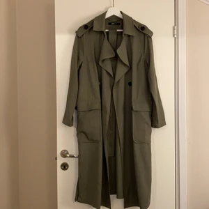 Trenchcoat från Gina Tricot  - Supersnygg militärgrön trenchcoat från Gina Tricot. Säljer pga att jag inte använder den. Använd ytterst få gånger. Midjeband tillkommer, dock har ena hållaren till bandet gått sönder på högra sidan. Storlek 38, säljer för 70kr + frakt.
