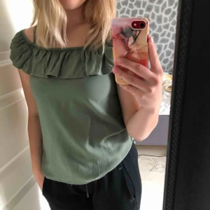  - En militärgrön, lite finare T-shirt. Har bara använts ett par gånger. Den är ganska tunn och tyget är ganska ”fladdrigt”. Den har en offsholder volang och axelband som ej går att justeras.  