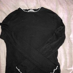  - Svart ribbad långärmad T-shirt med vita volanger i kragen och slutet av ärmen, köpt på Zara. 
