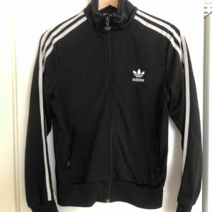  - Svart Adidas zip-tröja i wct-material, vit stor logga på ryggen och mindre logga på ena bröstet .
