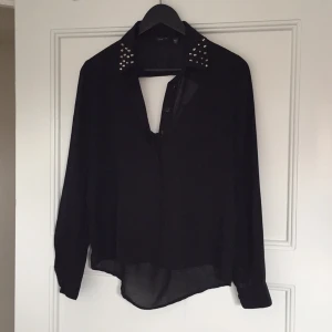  - Svart blus med nitar på kragen. Öppen rygg bak (se bild) samt knäppning fram. Köpt i New York.