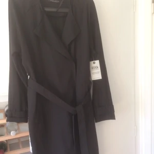  - Knälång, svart kappa, tunt tyg som passar till sommar och vår. Dolda knappar och band i midjan. Jättefin, säljer då jag fått en nästan likadan till. Den är aldrig använd. Det står strl S i den men den är mer som en M i storlek. 