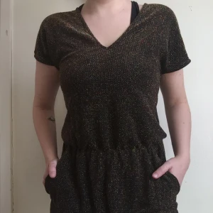  - V-ringad jumpsuit i guld och svart jersey. Oerhört bekväm, dock aldrig använd! Har fickor fram, avsmalnade ben och knäpps med en knapp i ryggen.   Jag är 165 cm lång och på mig är benen pösiga, men inte för långa och midjan sitter strax ovanför naveln.  