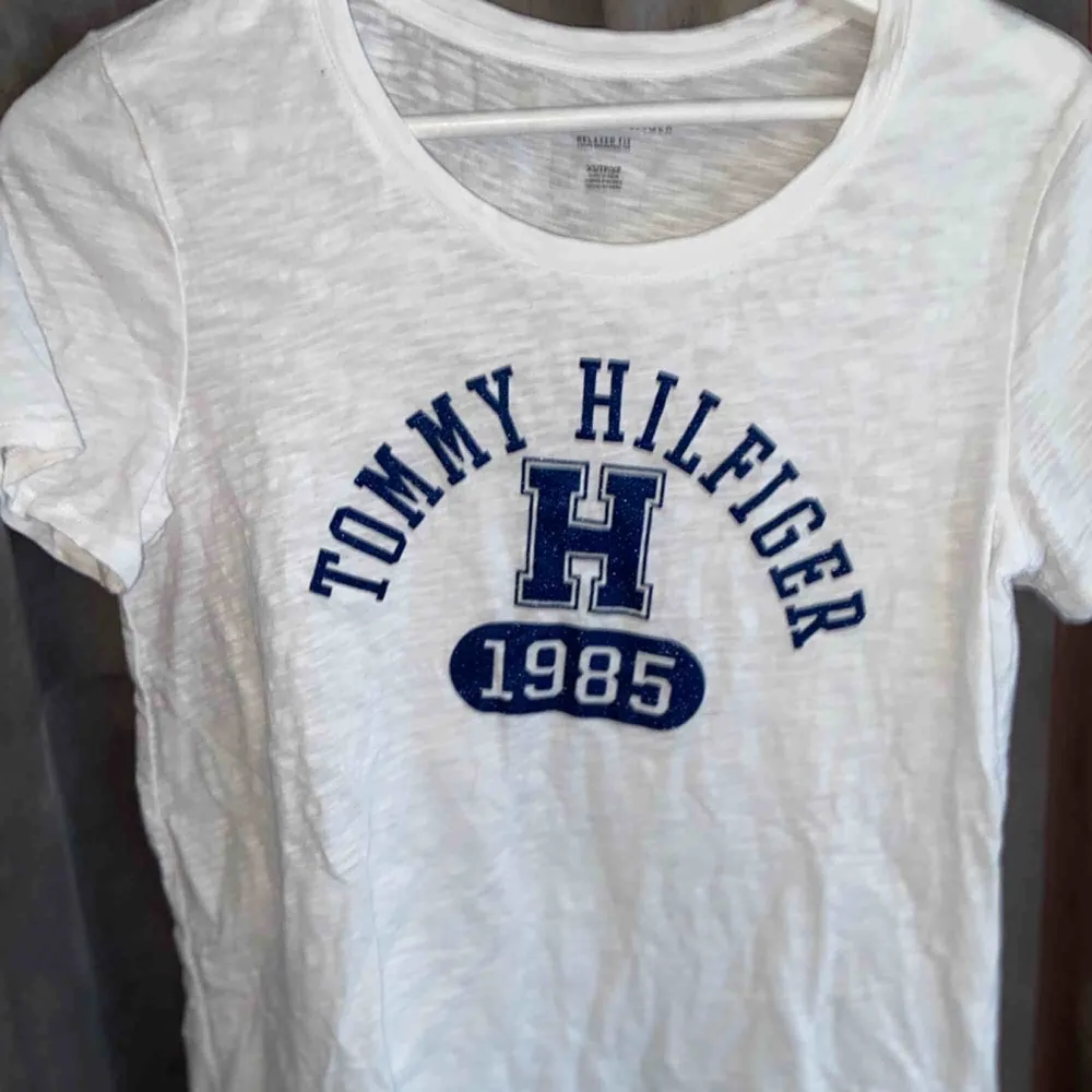 Vit Tommy Hilfiger t-shirt med blått tryck på bröstet. Denna är aldrig använd och köpt i höstas. Superfin och bra till sommaren.. T-shirts.