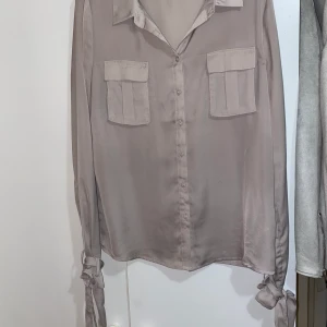 Skjortblus från NAKD - En skjortblus i en greige (beige/grå) färg. Hyfsat tunn. Den har knytband runt handlederna. Från Rut&Circle men köpt på nakd. Passar även xs.
