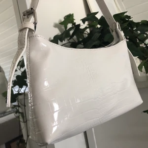 Vit väska från shein - Vit väska med krokodilmönster. Väskan är helt ny, aldrig använd. Säljer pga har redan en liknande. Väskan har justerbara band. Passar perfekt att ha under armen. Ca 25cm på lång. 