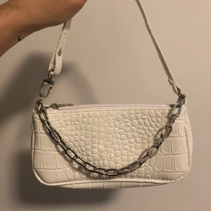 Vit handväska - Säljer en fin vit handväska med snyggt kedjesmycke som går att ta av om man vill! Den är ca 12 cm i höjd och 21cm i bredd