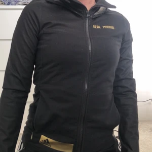 Kofta Adidas stl xs - En svart kofta från adidas med texten ”real madrid”, gulddetaljer och Luva. Stl xs