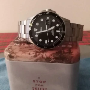 Herrklocka: Fossil Diver  - Märke:Fossil Diver  Pris:500 kr Diameter :42 mm Tjocklek:12 mm Armbandsmaterial: Rostfri stål  Armbandsbredd: 22 mm Glas: Mineral/Safir-mix Urverk: Quartz Vattenskydd:10 atm (bad/simning) Datumvisning:Ja Typ av spänne:Viklås Armbandsfärg:Silver Boettmaterial: Rostfritt stål Boettfärg: Silver Urskivefärg:Svart Tyvärr så finns det ett märke mellan 15 å 30 som syns på bild nummer ett