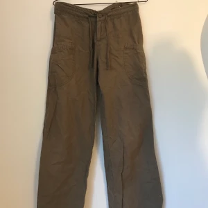 Bruna utsvängda byxor - Lågmidjade bruna, nästan gröna, byxor. Två fickor fram och möjlighet att dra ihop utsvängnigen med hjälp av snören längst ner på benen. Byxorna liknar ”cargo-pants”. Storleken skulle jag säga är 34-36. De är i mycket gott skick!