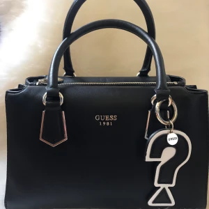 Guess handväska - Guess handväska med guld detaljer. Dustbag och snyggt axelband medföljer. Väskan har tre fack med ytterligare 4 små fickor på insidan. Använd ett par gånger men ser helt ny ut. Kan mötas i hela Stockholmsområdet. Original pris är 1600kr