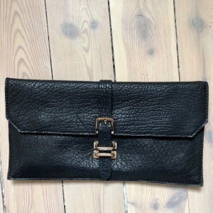  - Svart clutch från Tiger of Sweden med guldfärgat spänne och guldtryck baktill med loggan. Ett stor fack med en innerficka med dragkedja. Nyskick