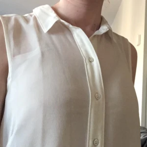  - Vit blus i tunn polyester. Rak, vid passform. Ärmlös med knappar fram och liten krage. Längre bak än fram. 

Jag skickar med posten eller möter upp i Stockholm. Betalning via Plick, kontoinbetalning, kontant eller swish.

