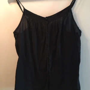  - Svart tunt linne med smala axelband. Knappar fram. Justerbara axelband. 