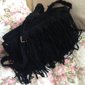  - Svart fransväska i mockaimitation. Axelremmen är flätad och har guldigt spänne.

Bredd: 34 cm
Höjd: 22 cm
Djup (botten): 11 cm