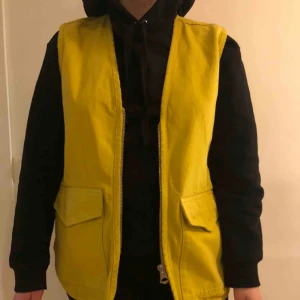  - Senapsgul väst i jeans med små fickor på framsidan och en stor ficka på baksidan. Dragkedja fram och på fickan 