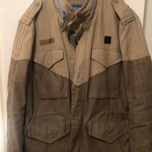 Adidas Originals ST M65 military sheep skin lined khaki vinterjacka - Jackan är i storlek L och är bra skick förutom två små hål. Ett vid knappen på fickan på höger sida och ett strax ovanför fickan på vänster sida. Finns även en liten fläck som knappt är synlig längs ner på jackan.